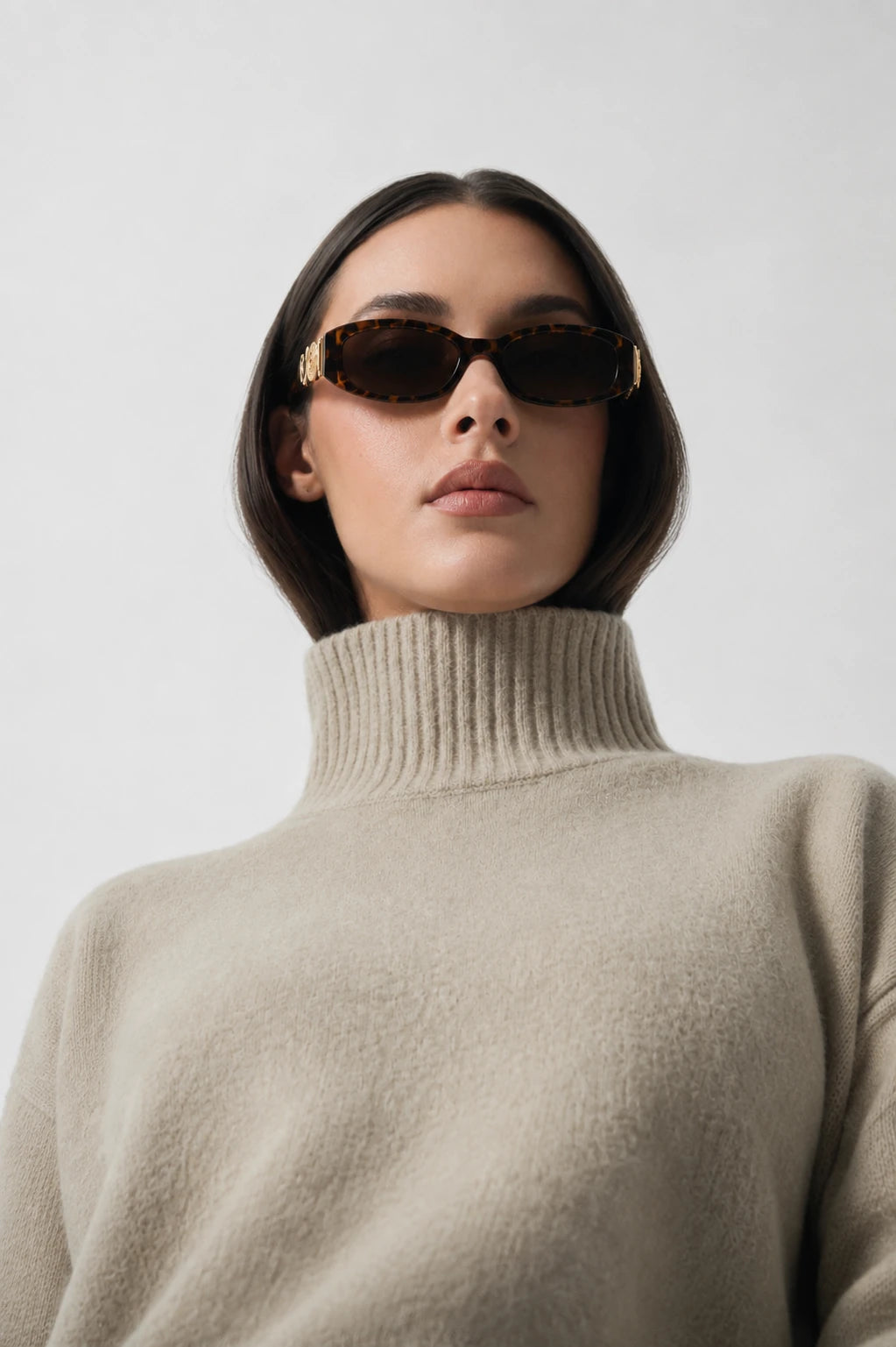 A model with short dark hair is shown from the chest up wearing a light beige ribbed turtleneck sweater and dark brown sunglasses with gold metal frames, set against a plain light gray background.