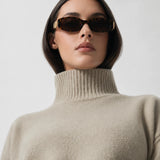 A model with short dark hair is shown from the chest up wearing a light beige ribbed turtleneck sweater and dark brown sunglasses with gold metal frames, set against a plain light gray background.