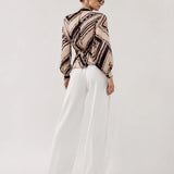 A woman is shown from the back wearing a beige and black patterned long-sleeve blouse tucked into white wide-leg trousers, set against a plain light gray studio background.