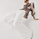 A model wearing a beige and black patterned long-sleeve top, white wide-leg trousers, and dark pointed shoes poses on a seamless white studio background.