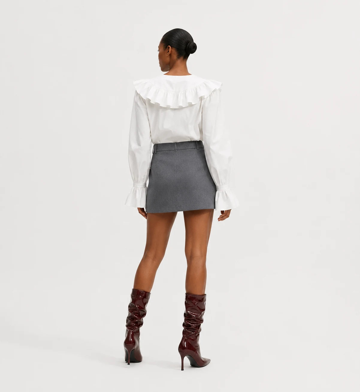 A model shown from the back wearing a white long-sleeve blouse with a large ruffled collar, a gray mini skirt, and dark burgundy knee-high boots, set against a plain light gray studio background.