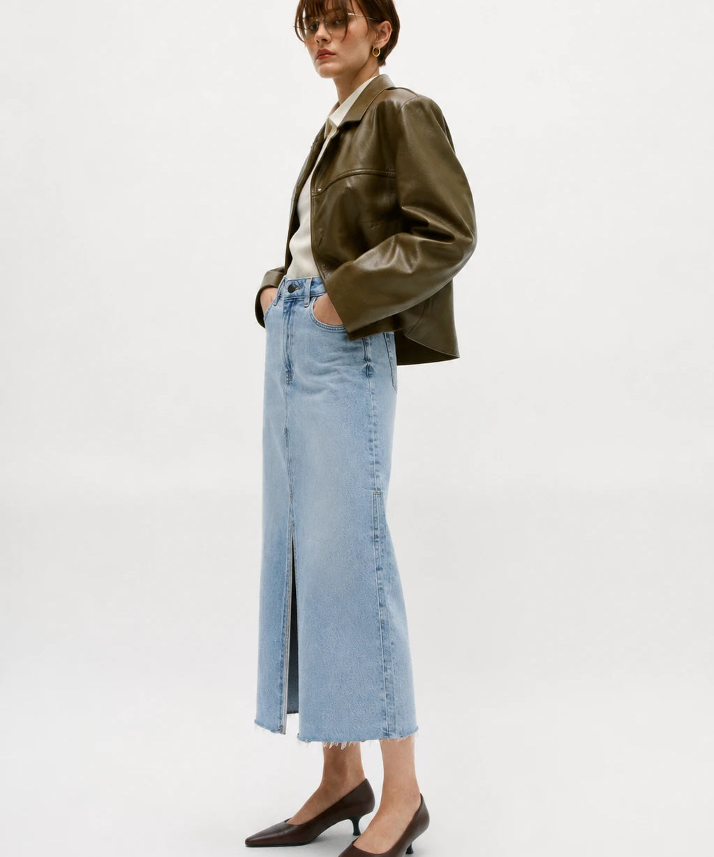 A model stands in profile wearing a dark olive green leather jacket, a light blue high-waisted midi skirt with a front slit, a cream shirt, and dark brown pointed-toe flats against a plain white studio background.