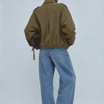 A model shown from behind wearing an olive green cropped utility jacket, wide-leg light blue denim jeans, and black pointed-toe heels, holding a dark brown textured shoulder bag against a light gray studio background.