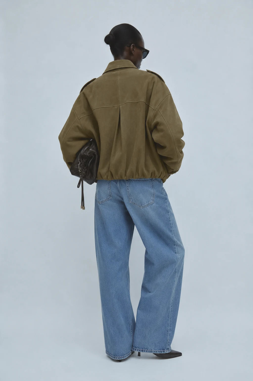 A model shown from behind wearing an olive green cropped utility jacket, wide-leg light blue denim jeans, and black pointed-toe heels, holding a dark brown textured shoulder bag against a light gray studio background.