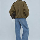 A model shown from behind wearing an olive green cropped utility jacket, wide-leg light blue denim jeans, and black pointed-toe heels, holding a dark brown textured shoulder bag against a light gray studio background.
