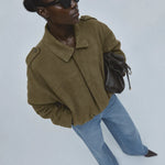 A model wearing an olive green cropped jacket, light blue jeans, black sunglasses, and carrying a dark brown leather bag, photographed from a high angle against a light gray background.