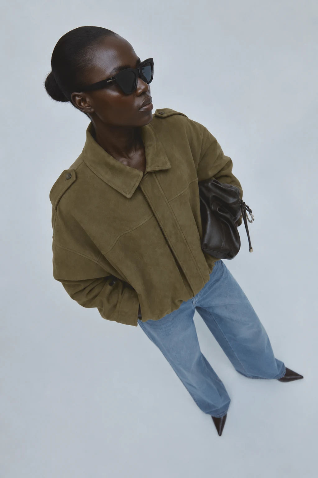 A model wearing an olive green cropped jacket, light blue jeans, black sunglasses, and carrying a dark brown leather bag, photographed from a high angle against a light gray background.