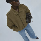 A model wearing an olive green cropped jacket, light blue jeans, black sunglasses, and carrying a dark brown leather bag, photographed from a high angle against a light gray background.