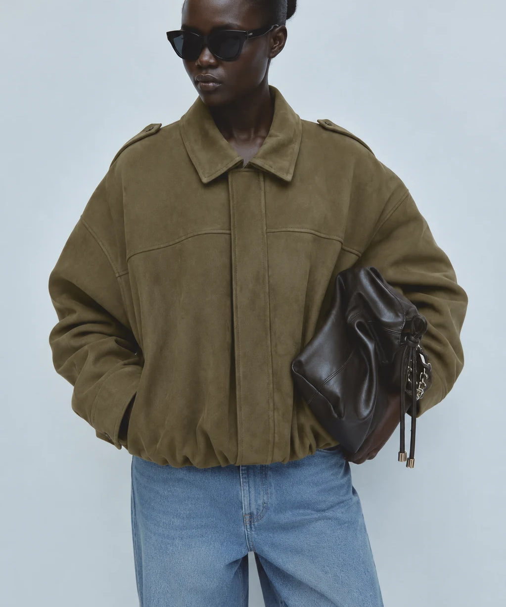A model wearing an oversized olive green suede jacket, blue jeans, black sunglasses, and holding a dark brown leather bag against a light gray studio background.