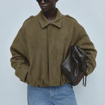 A model wearing an oversized olive green suede jacket, blue jeans, black sunglasses, and holding a dark brown leather bag against a light gray studio background.