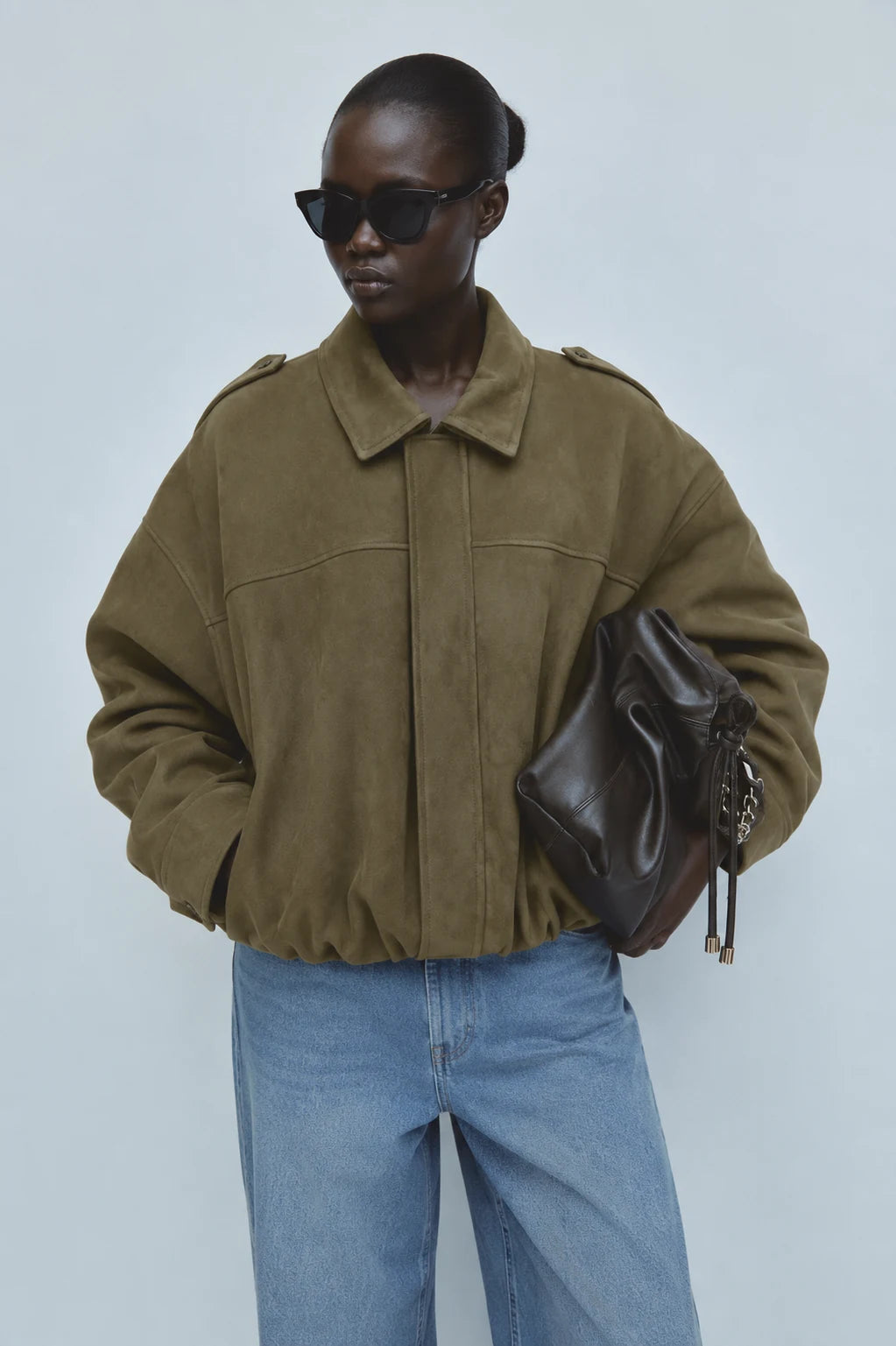 A model wearing an oversized olive green suede jacket, blue jeans, black sunglasses, and holding a dark brown leather bag against a light gray studio background.