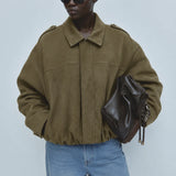A model wearing an oversized olive green suede jacket, blue jeans, black sunglasses, and holding a dark brown leather bag against a light gray studio background.