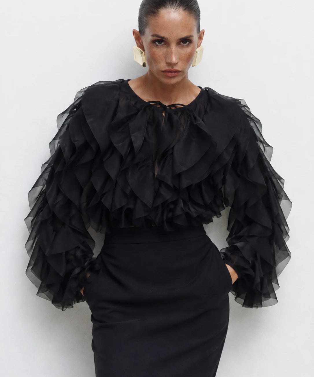 A model wearing a black ruffled long-sleeve blouse with sheer layered sleeves and a black pencil skirt, standing against a light gray studio background.