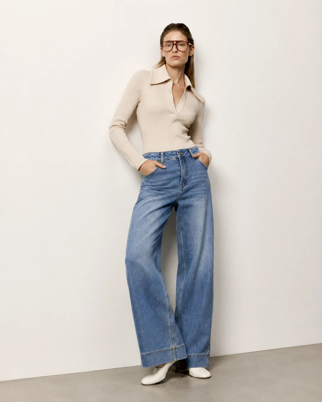 A woman wearing a beige long-sleeve collared knit top and high-waisted, wide-leg blue denim jeans, standing against a plain off-white wall.