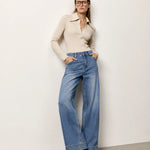 A woman wearing a beige long-sleeve collared knit top and high-waisted, wide-leg blue denim jeans, standing against a plain off-white wall.