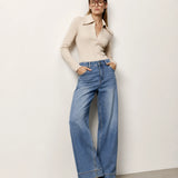 A woman wearing a beige long-sleeve collared knit top and high-waisted, wide-leg blue denim jeans, standing against a plain off-white wall.