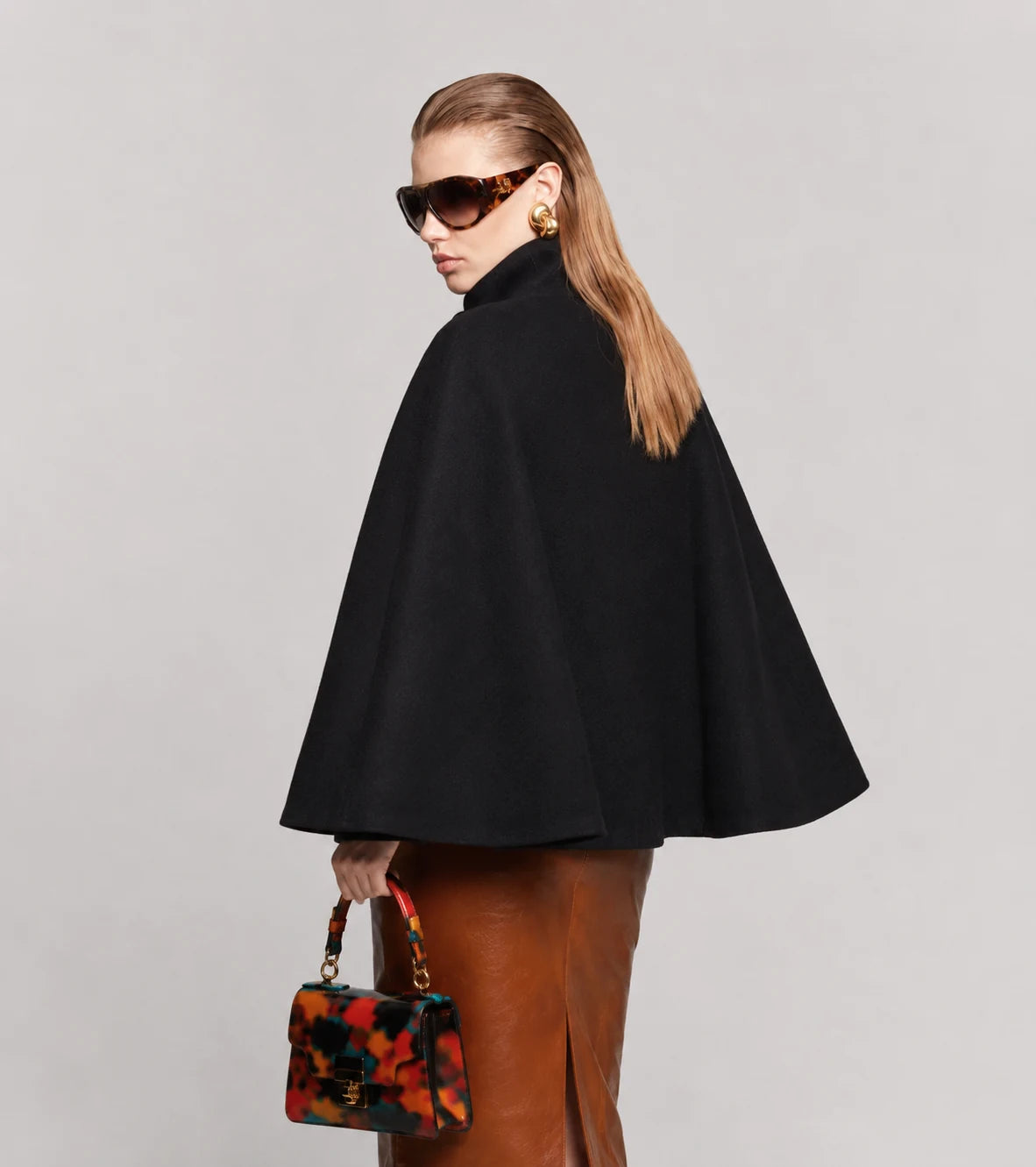 A woman modeling a black cape-style coat with a high mock neck, shown from the back and side against a light gray studio background. She is wearing brown leather pants and holding a small floral handbag.