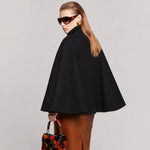 A woman modeling a black cape-style coat with a high mock neck, shown from the back and side against a light gray studio background. She is wearing brown leather pants and holding a small floral handbag.