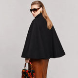 A woman modeling a black cape-style coat with a high mock neck, shown from the back and side against a light gray studio background. She is wearing brown leather pants and holding a small floral handbag.