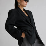 A model wearing a black oversized blazer, gray trousers, black sunglasses, and silver earrings, posed against a light gray studio background.
