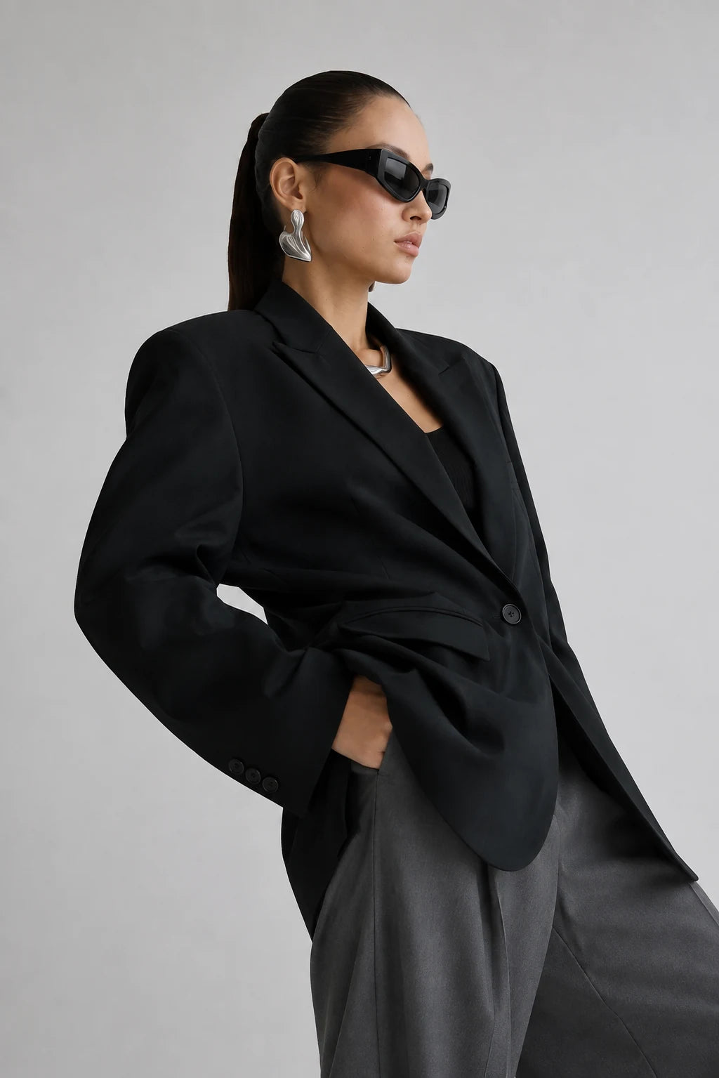 A model wearing a black oversized blazer, gray trousers, black sunglasses, and silver earrings, posed against a light gray studio background.