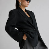 A model wearing a black oversized blazer, gray trousers, black sunglasses, and silver earrings, posed against a light gray studio background.