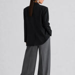 A model shown from the back wearing a black oversized blazer and wide-leg charcoal gray trousers, styled with black sunglasses, against a light gray studio background.