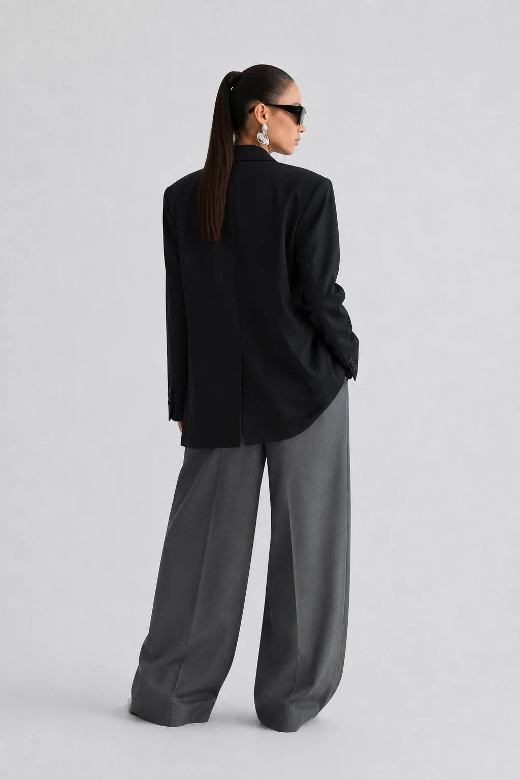 A model shown from the back wearing a black oversized blazer and wide-leg charcoal gray trousers, styled with black sunglasses, against a light gray studio background.