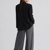 A model shown from the back wearing a black oversized blazer and wide-leg charcoal gray trousers, styled with black sunglasses, against a light gray studio background.