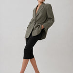 A model wearing an oversized olive green blazer, black capri leggings, and black pointed-toe heels, posing against a light gray studio background.