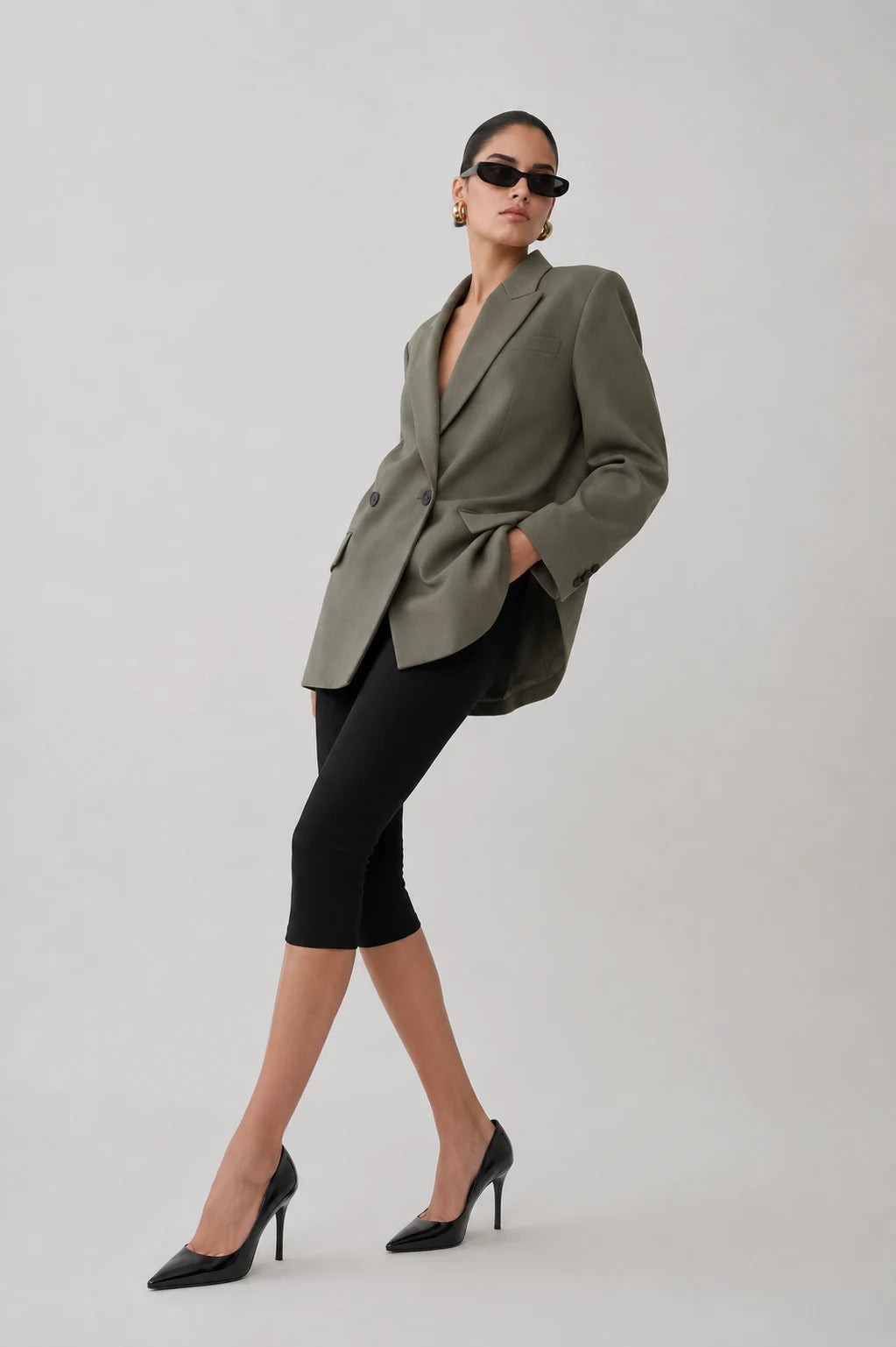 A model wearing an oversized olive green blazer, black capri leggings, and black pointed-toe heels, posing against a light gray studio background.