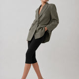 A model wearing an oversized olive green blazer, black capri leggings, and black pointed-toe heels, posing against a light gray studio background.