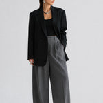 A model stands against a light gray studio background wearing a black blazer over a black top, paired with high-waisted, wide-leg gray trousers and black pointed shoes.