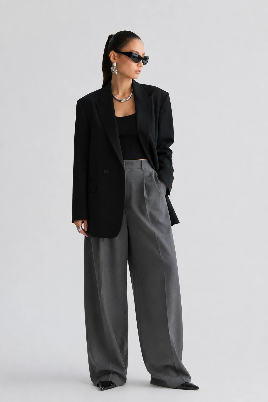 A model stands against a light gray studio background wearing a black blazer over a black top, paired with high-waisted, wide-leg gray trousers and black pointed shoes.