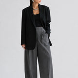 A model stands against a light gray studio background wearing a black blazer over a black top, paired with high-waisted, wide-leg gray trousers and black pointed shoes.