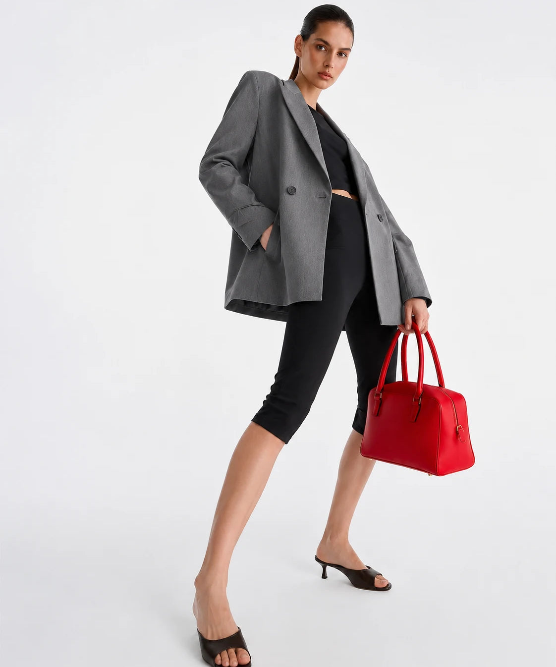 A model poses in a gray double-breasted blazer, black cropped leggings, and black slide sandals, holding a bright red structured handbag against a clean white studio background.