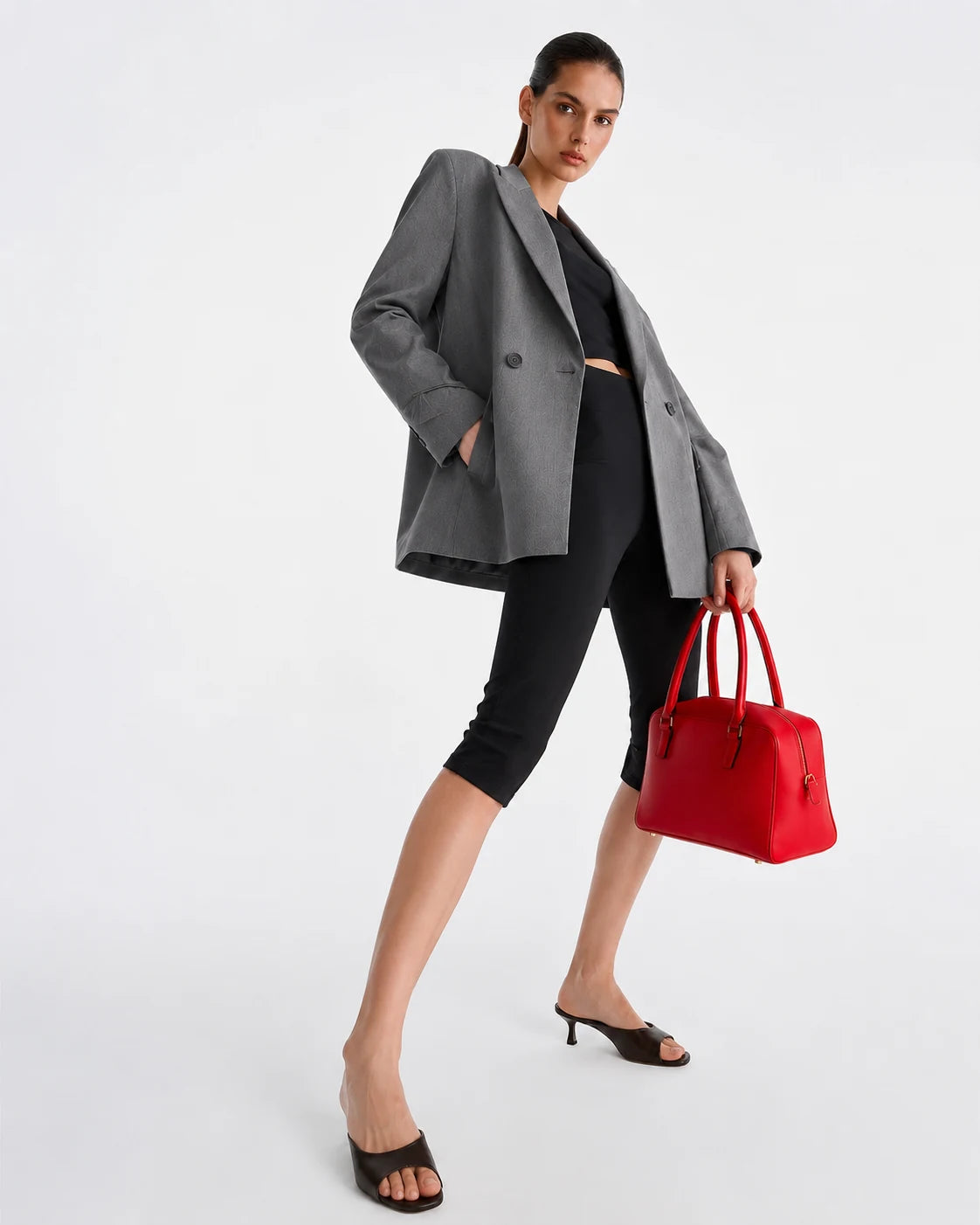 A model poses in a gray double-breasted blazer, black cropped leggings, and black slide sandals, holding a bright red structured handbag against a clean white studio background.