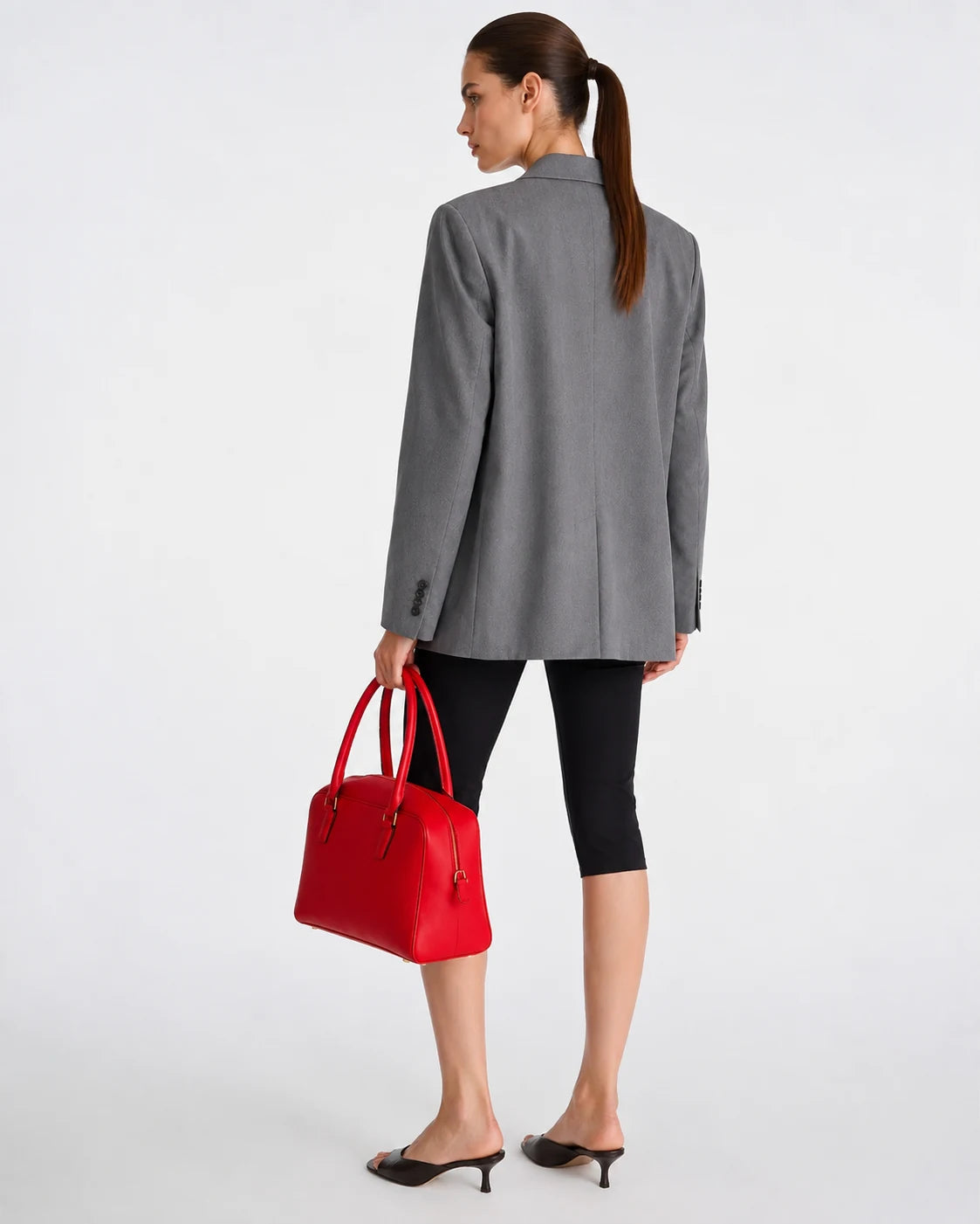 A woman shown from the back wearing a gray blazer, black cropped leggings, and black heeled mules, while holding a bright red structured handbag against a clean white studio background.