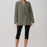 A model stands against a light gray studio background wearing an oversized olive green double-breasted blazer, black cropped leggings, and black pointed-toe heels.