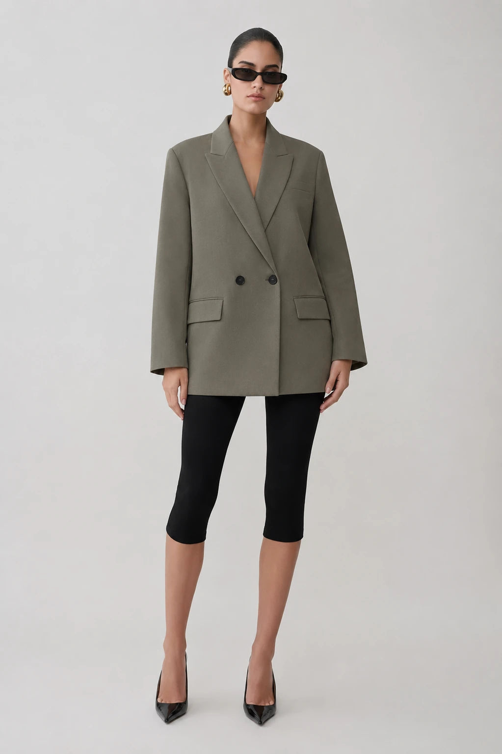 A model stands against a light gray studio background wearing an oversized olive green double-breasted blazer, black cropped leggings, and black pointed-toe heels.