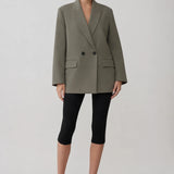 A model stands against a light gray studio background wearing an oversized olive green double-breasted blazer, black cropped leggings, and black pointed-toe heels.