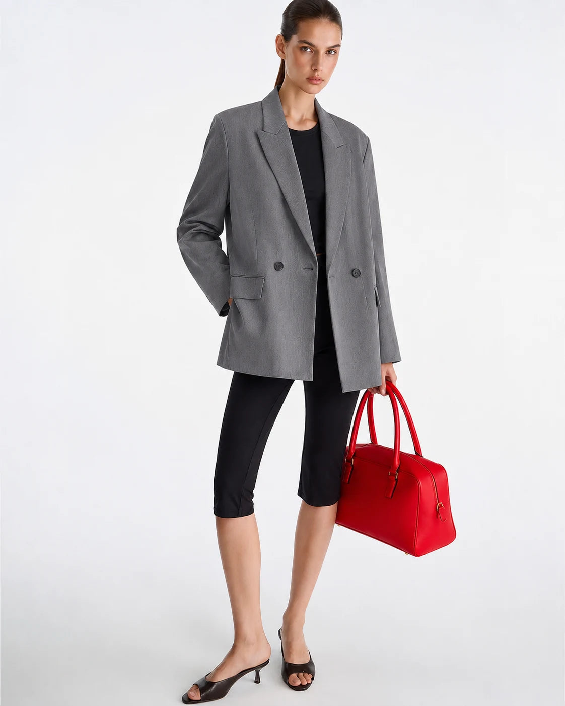 A model stands against a light gray studio background wearing an oversized gray blazer over a black top and black capri leggings, holding a bright red structured handbag.