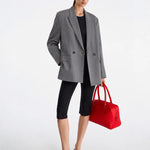 A model stands against a light gray studio background wearing an oversized gray blazer over a black top and black capri leggings, holding a bright red structured handbag.