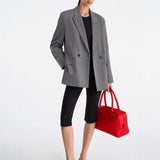 A model stands against a light gray studio background wearing an oversized gray blazer over a black top and black capri leggings, holding a bright red structured handbag.