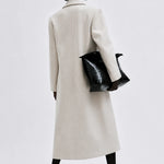 A model shown from the back wearing a long, straight-line cream-colored coat, holding a black textured handbag, and wearing black pointed-toe heels against a light gray studio background.