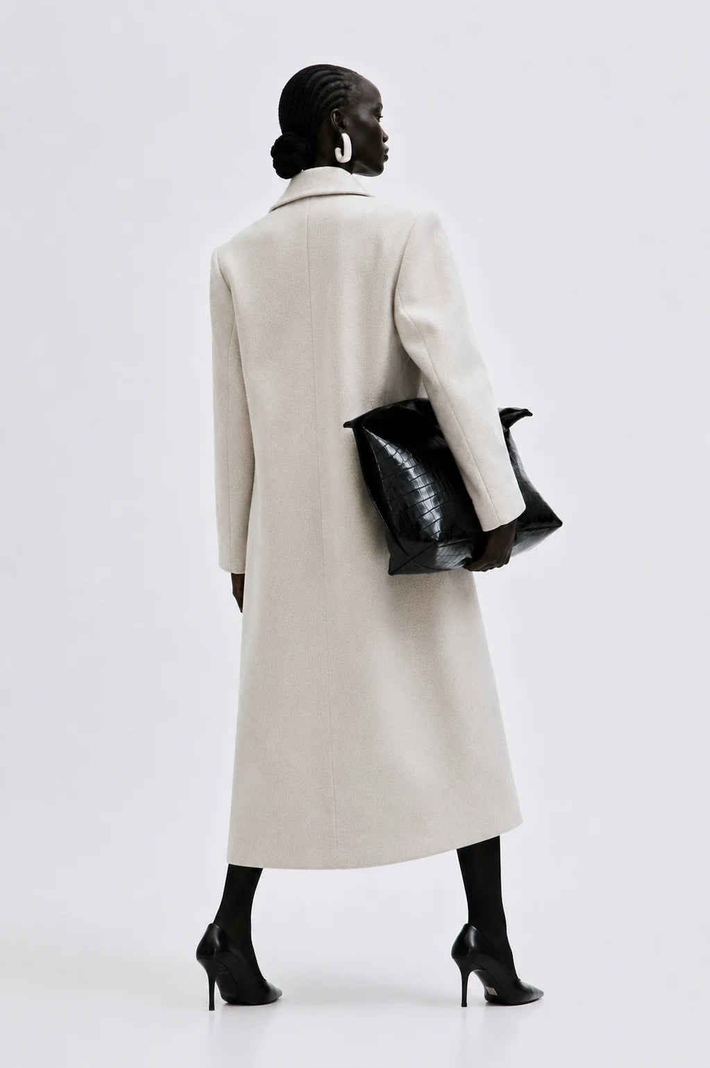 A model shown from the back wearing a long, straight-line cream-colored coat, holding a black textured handbag, and wearing black pointed-toe heels against a light gray studio background.