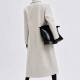 A model shown from the back wearing a long, straight-line cream-colored coat, holding a black textured handbag, and wearing black pointed-toe heels against a light gray studio background.