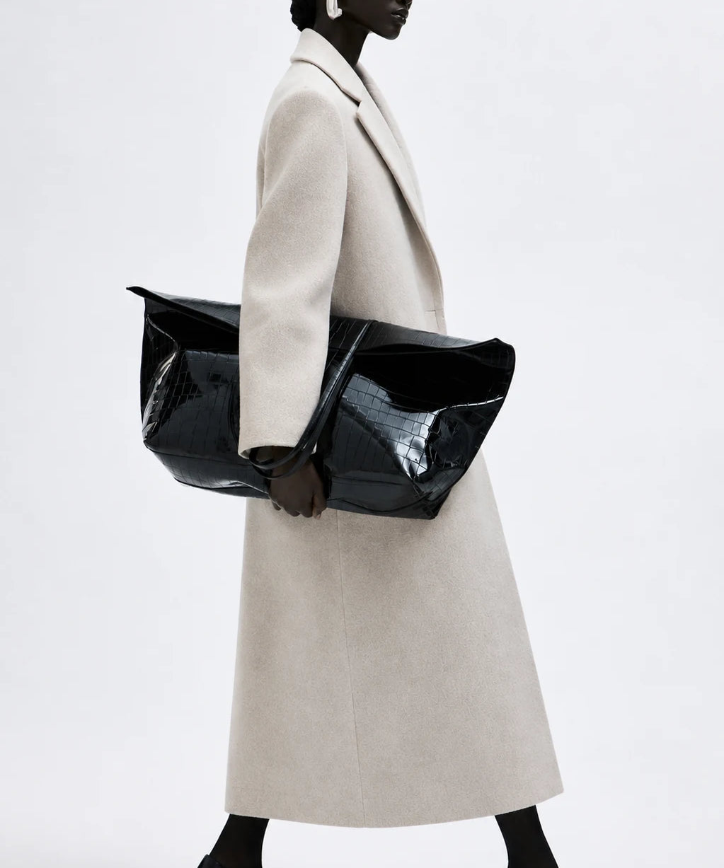 kvinna bär beige Seraphina Coat med svarta kalcksor, väska och stora silver örhängen mot en vit vägg