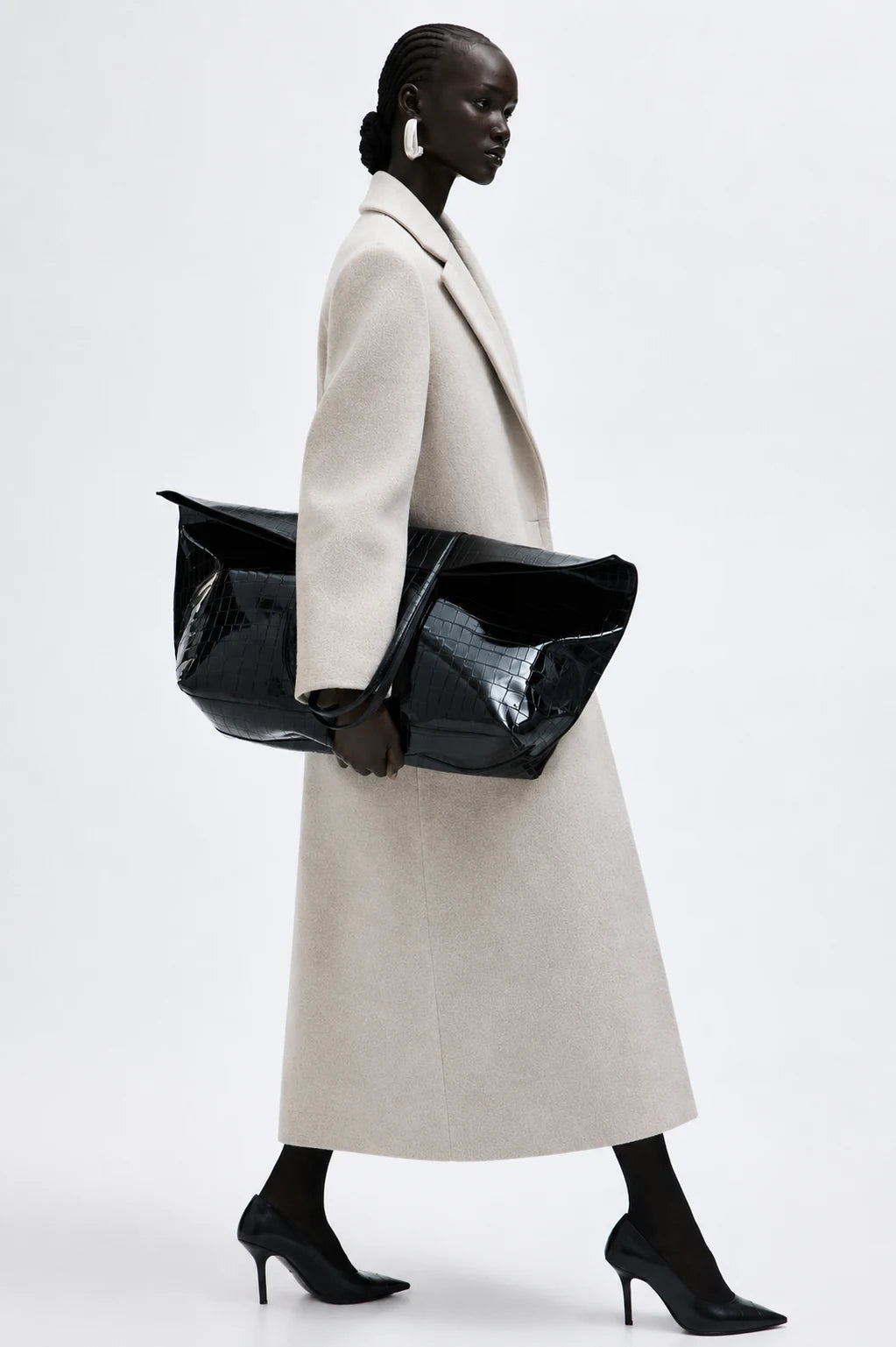 kvinna bär beige Seraphina Coat med svarta kalcksor, väska och stora silver örhängen mot en vit vägg
