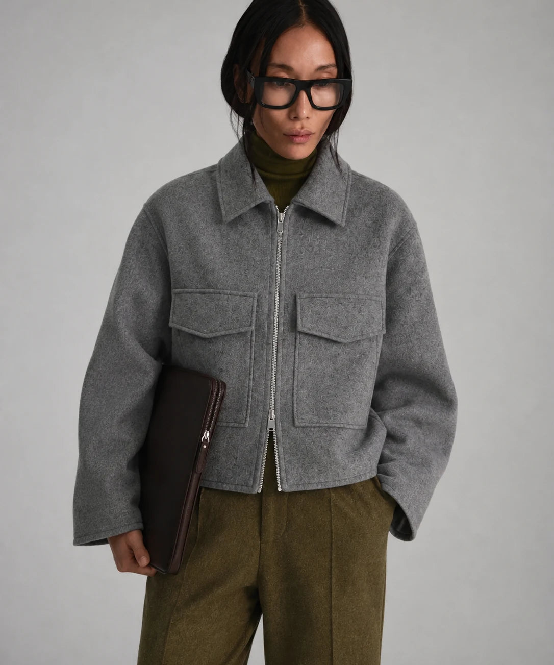 A model wearing a cropped gray wool-blend jacket with a front zipper and flap pockets, styled over an olive green turtleneck and olive trousers, holding a dark brown clutch against a neutral gray background.